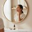 A woman with medium-deep skin tone applies Derma Made Melafade Serum to her cheek in front of a mirror. The 4-inch serum bottle sits on a marble counter beside a ceramic dish and white flower in a softly lit, minimal skincare setup.